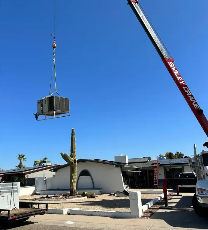A crane lifts a new rooftop heating and cooling unit over a single-story Arizona home with a saguaro cactus in the front yard.