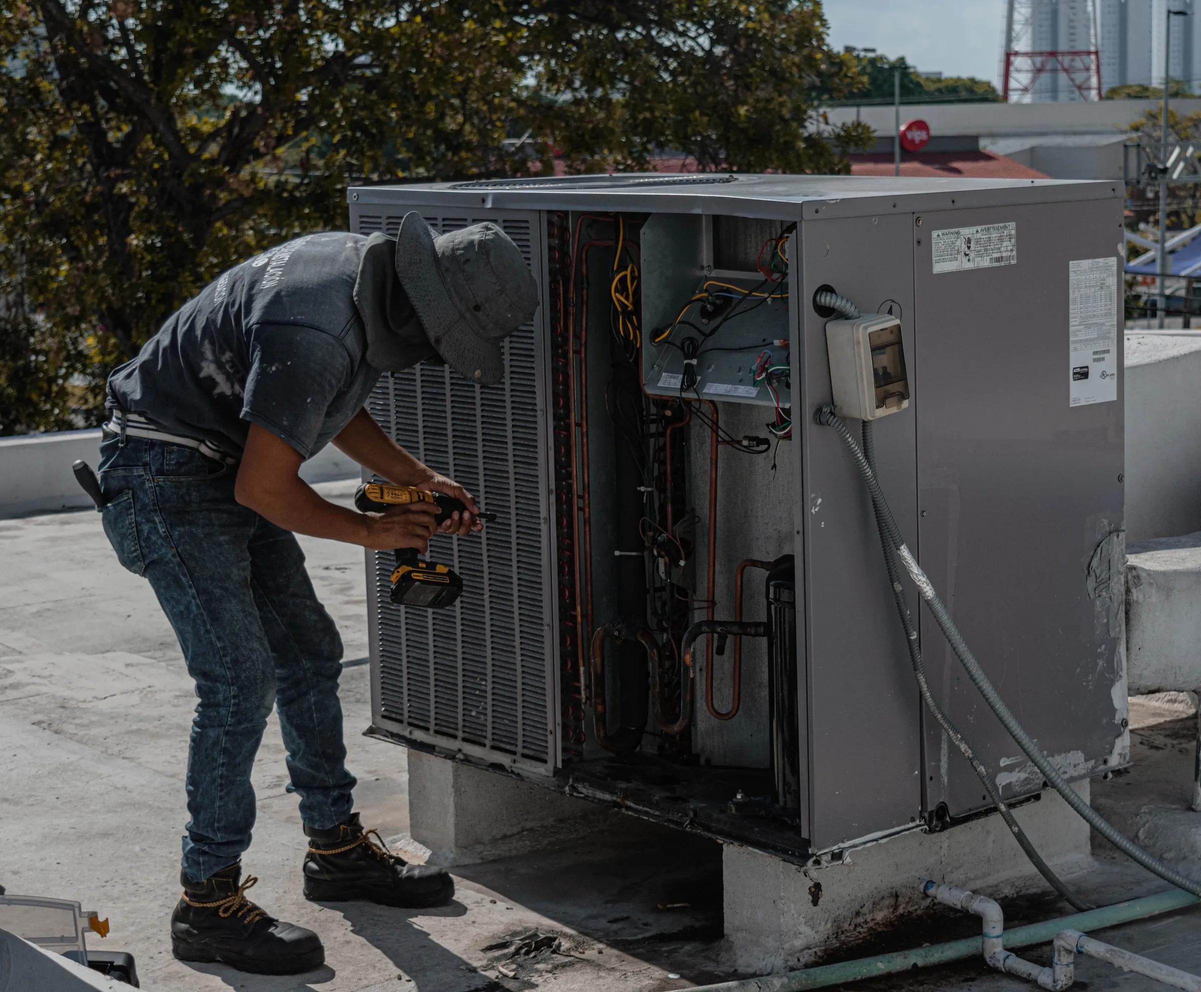A technician uses a cordless drill on an open rooftop heating and cooling unit.