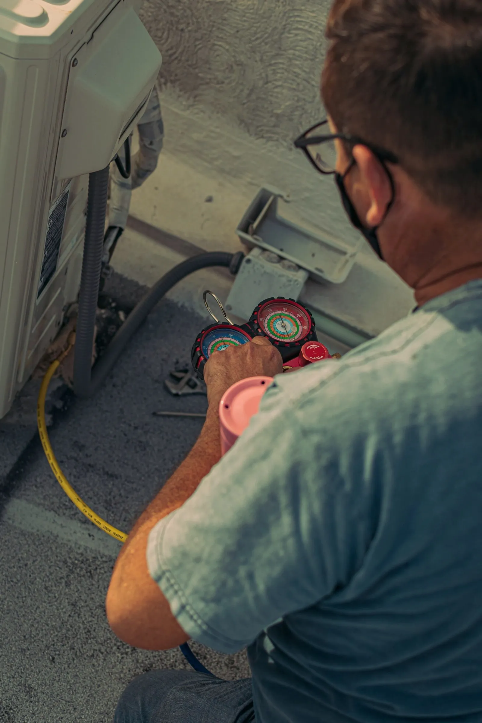 A technician holds a refrigerant gauge set while checking an outdoor air conditioning unit.