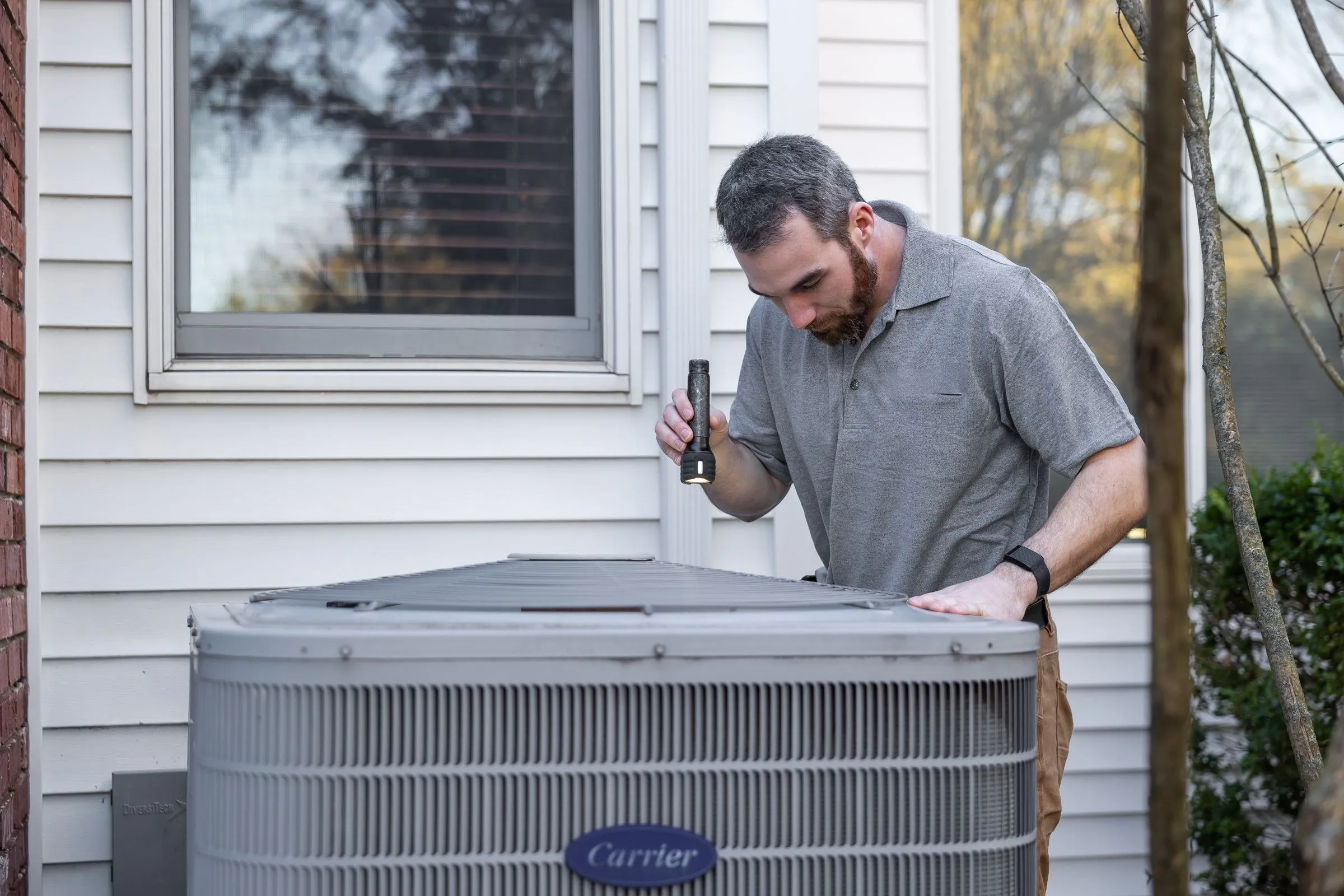 A technician uses a flashlight to look inside an outdoor air conditioning unit beside a house.